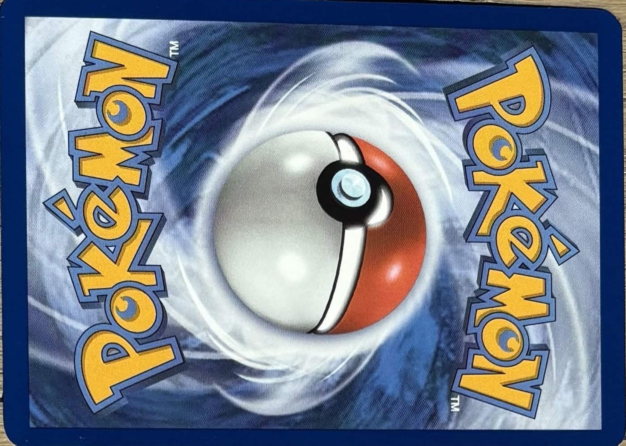 The back of a genuine English Pokémon card, photographed sideways on a wooden table: dark blue border, white swirl around the Poké Ball, the Pokémon logo twice with a trademark sign.