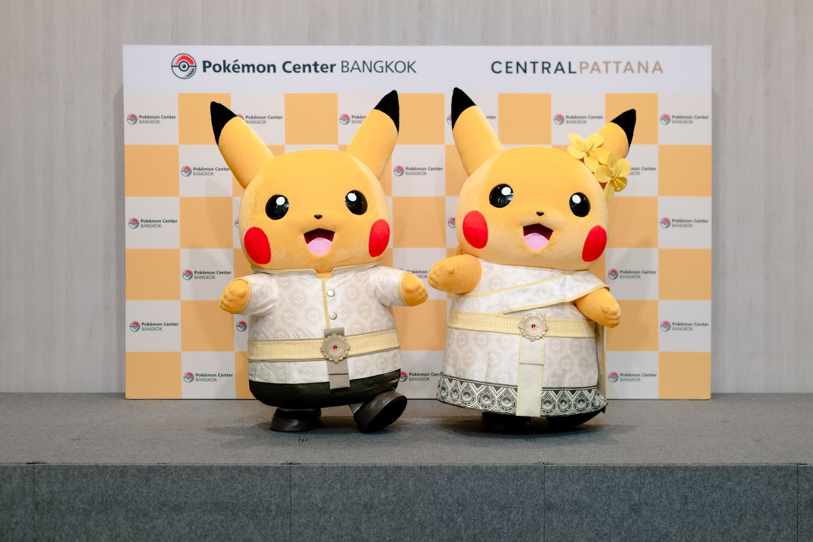 Two costumed walking Pikachu in cream Thai dress on a stage, in front of a Pokémon Center BANGKOK and Central Pattana backdrop.