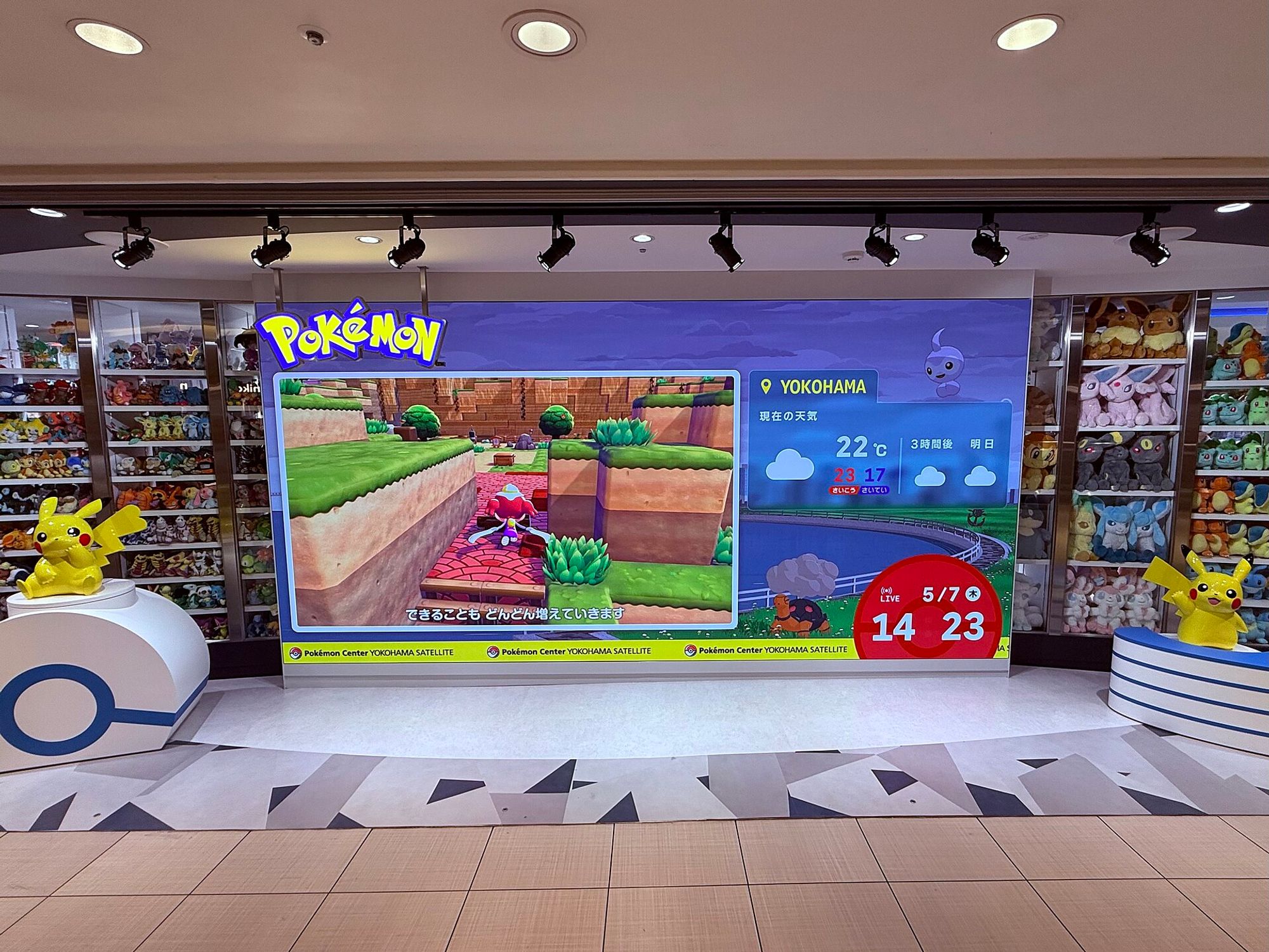 The storefront of Pokémon Center YOKOHAMA Satellite, a wall-sized screen between shelves of plush, with a Pikachu statue on each side.