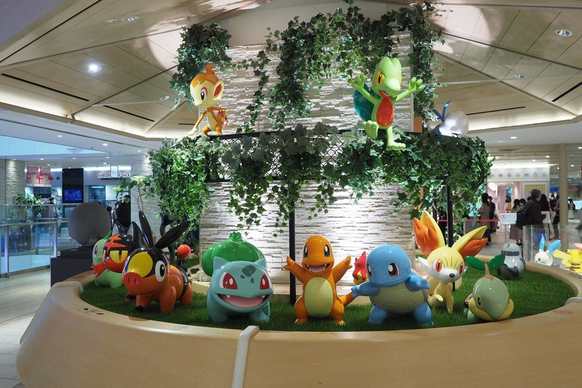 A garden display in a shopping mall concourse: life-size statues of Bulbasaur, Charmander, Squirtle, Tepig, Fennekin and Turtwig on artificial grass, with Chimchar and Treecko above on a vine-covered trellis.