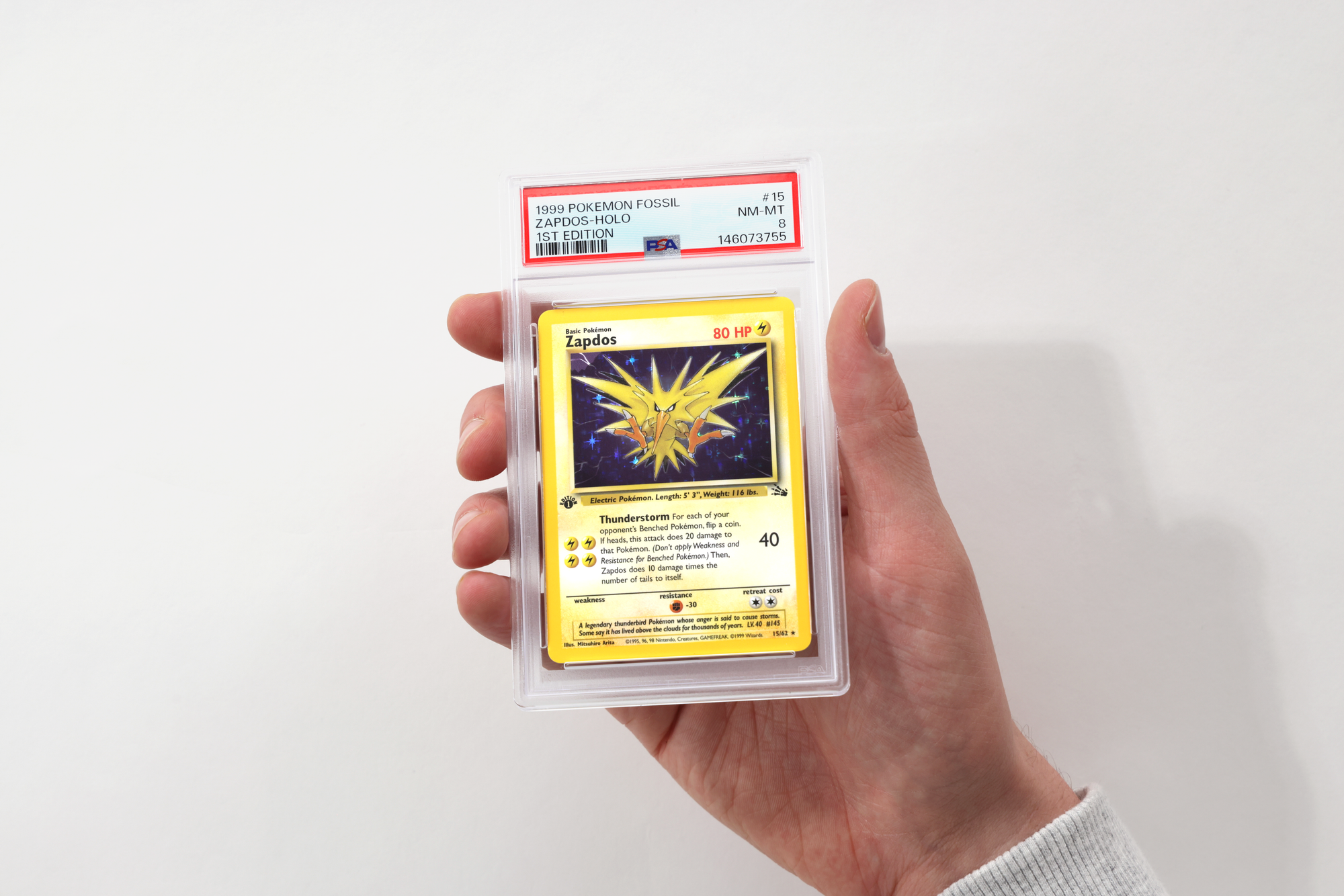 A hand holding a PSA holder containing a 1999 Pokemon Fossil first edition Zapdos holo graded NM-MT 8, with the certification number on the label.